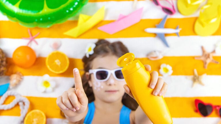 Young girl wearing sunglasses on beach towel reaching out with sunscreen in hands