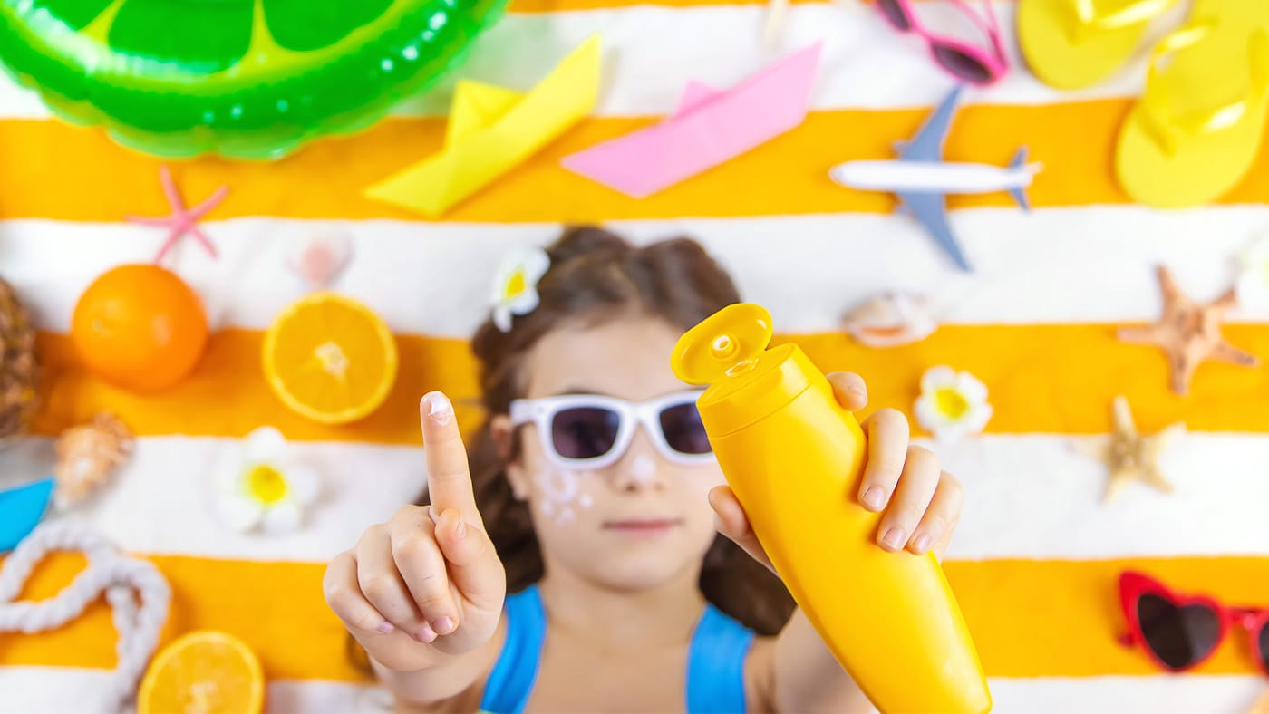 Young girl wearing sunglasses on beach towel reaching out with sunscreen in hands