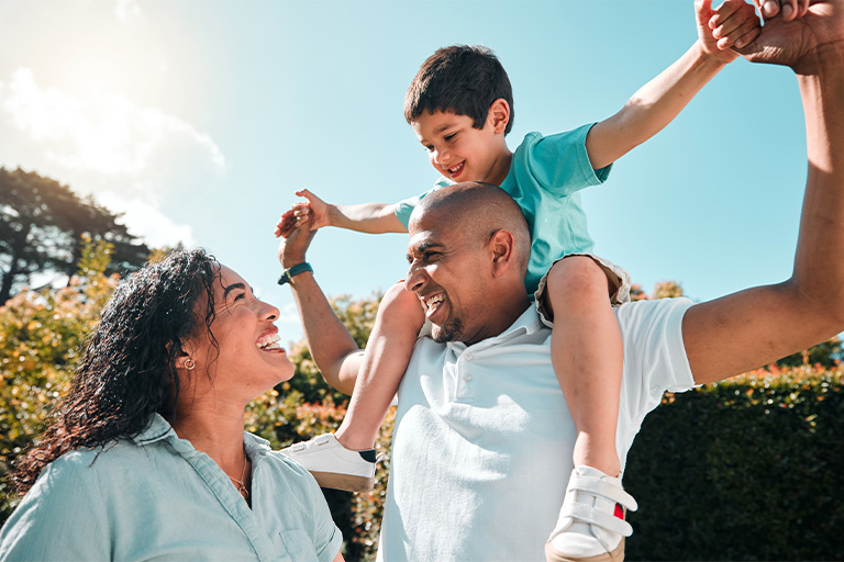 Married couple laughing together outside with their son on the father's shoulders