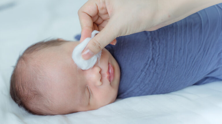 A newborn sleeping while his mother cleans his eye with a cotton pad