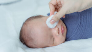 A newborn sleeping while his mother cleans his eye with a cotton pad