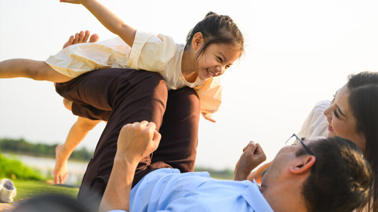 A family outdoors lying down, the father is lifting his daughter up with his legs.